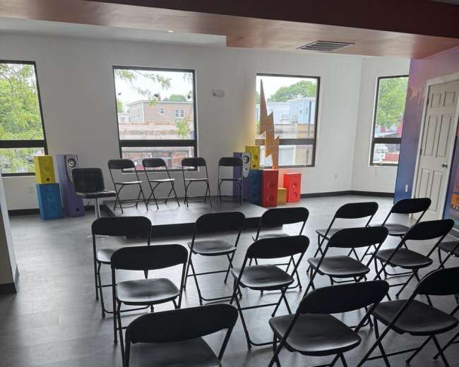 The image shows a room with several rows of black folding chairs arranged in front of a table and colorful speakers against the wall.