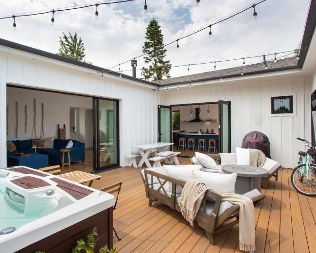 The image shows a modern outdoor patio featuring a hot tub, seating area, and a dining table, all surrounded by wooden decking and framed by white walls and string lights overhead.