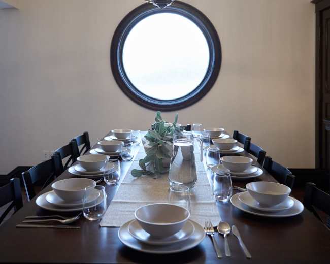 A long dining table is set with plates, glasses, and cutlery underneath a large round chandelier and a circular window.