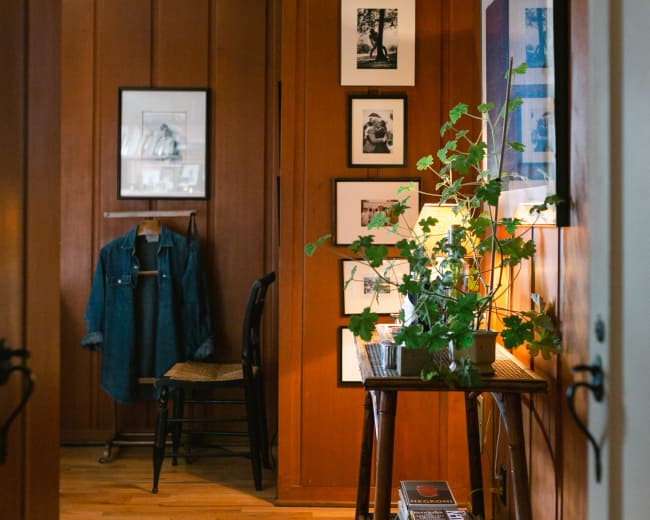 A hallway features wooden paneled walls, a small table with plants and books, and framed photographs hanging nearby.