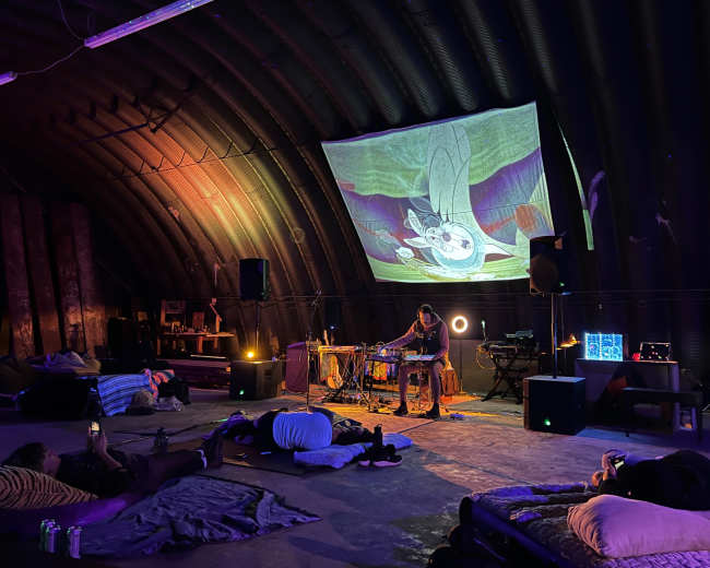 A musician performs on a small stage with a projector display in a dimly lit, spacious indoor venue where people relax on floor cushions.