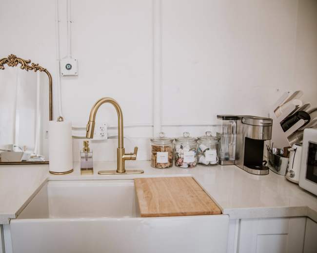 A kitchen countertop features a sink, gold faucet, mirror, and various kitchen appliances and jars.