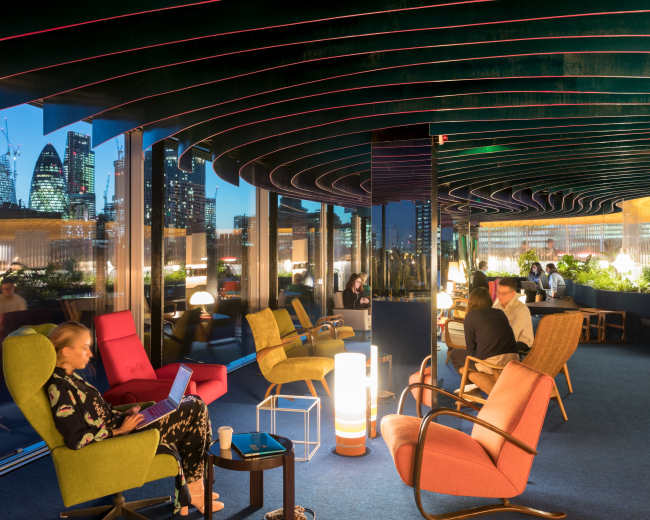 A modern lounge features colorful seating arrangements with large windows overlooking a city skyline at dusk.