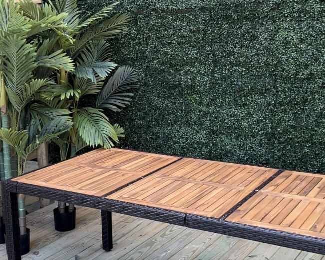 The image features a wooden table in front of a green artificial hedge and potted plants on a wooden deck.