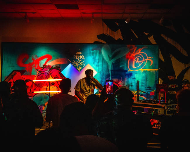 A musician performs on stage in front of a crowd, with vibrant graffiti artwork displayed behind them.