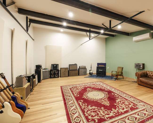 A spacious music rehearsal room with wooden floors, a large rug, an array of amplifiers, and a brown leather couch.