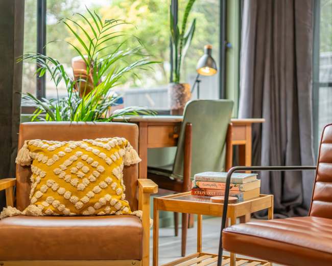 A cozy living space with two armchairs, a small wooden table with stacked books, and a desk in the background, surrounded by large windows and indoor plants.