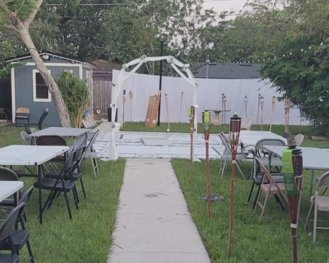 The image shows a pathway leading to a tent set up in a grassy area, flanked by chairs and tiki torches.