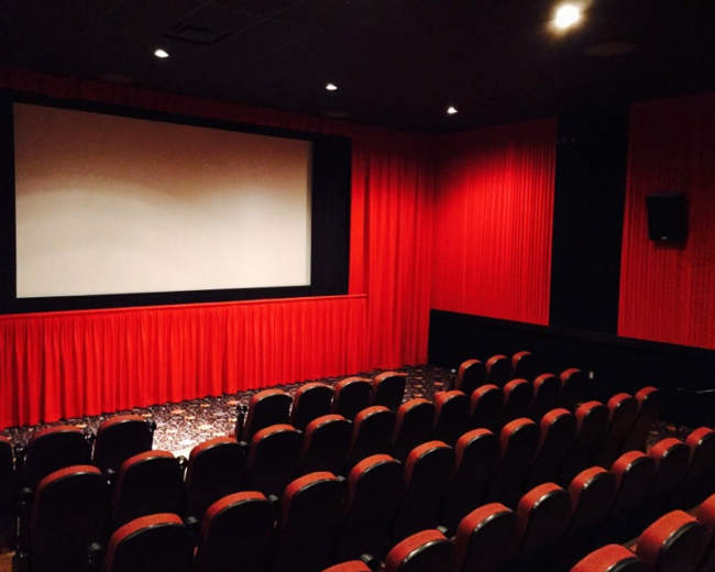 A small theater features red curtains, a large screen, and rows of empty red-upholstered seats.