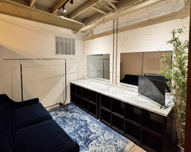 The image shows a spacious and minimalist studio with a dark blue couch, a wooden table, two mirrors, and overhead lighting against exposed brick walls.