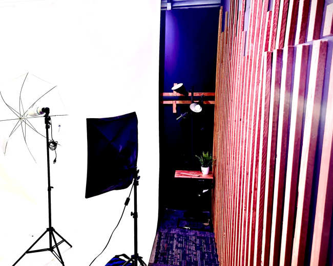 The image shows a photography studio setup with a white backdrop, two lighting stands, a reflector, and wooden paneling on one side.