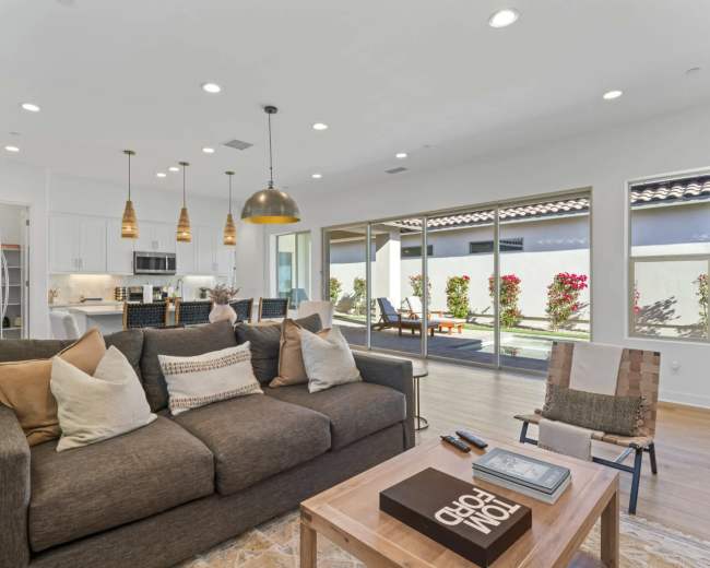 The image shows a modern living room with a large brown sofa, a coffee table, and glass doors that open to a patio area with a garden.