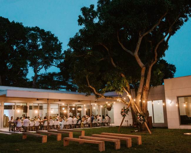 A large group of people dining outdoors under string lights next to a modern building surrounded by trees.