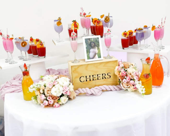 A table is adorned with colorful cocktails in elegant glasses, a decorative "CHEERS" sign, a framed photo, and floral arrangements.