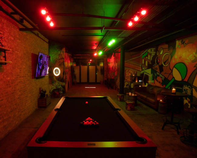 The image shows a dimly lit lounge area featuring a pool table, illuminated by red and green lights, with vibrant murals on the walls and a television on one side.
