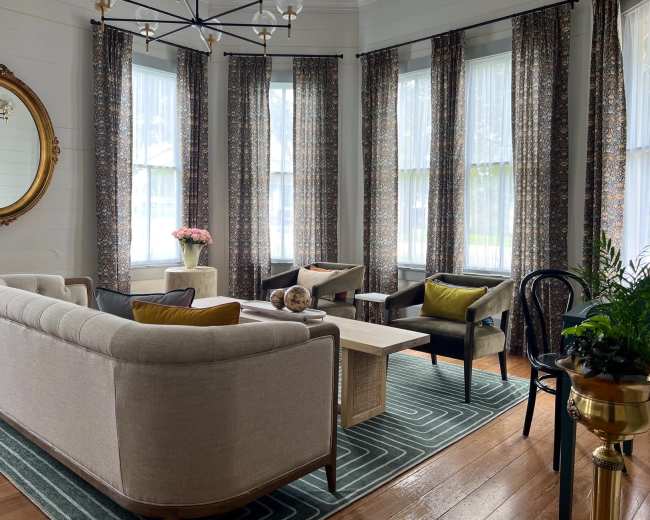 The image shows a brightly lit living room with large windows, featuring a mix of upholstered furniture, a coffee table, a decorative mirror, and potted plants.