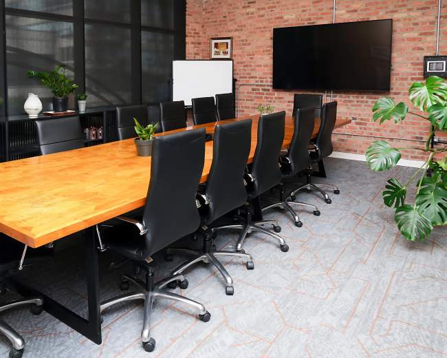 The image shows a modern conference room featuring a large wooden table surrounded by black ergonomic chairs, with a wall-mounted television and potted plants in the background.