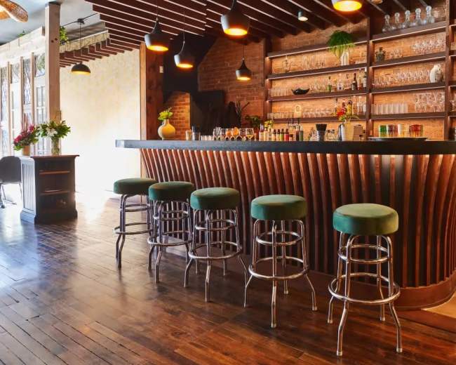 The image shows a bar with a curved wooden counter, adorned with shelves of glassware and decorative items, accompanied by several green upholstered stools.