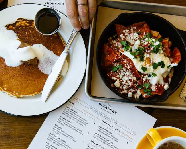 A hand with a knife and fork reaches for a plate of pancakes and a bowl of chilaquiles, accompanied by a coffee cup and a menu on a wooden table.