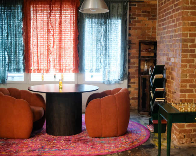 The image shows a cozy room with a round table surrounded by orange armchairs, brick walls, and large windows with colorful curtains, along with a nearby bookshelf and a small table featuring a chess set.