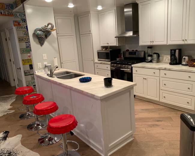 The image shows a modern kitchen with white cabinets, a marble countertop island with red stools, and stainless steel appliances.