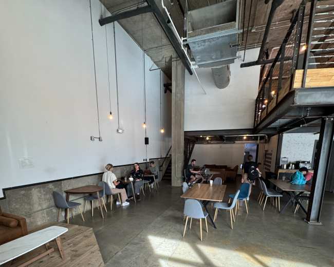 The image shows a spacious interior of a café or co-working space with tables and chairs arranged on a polished concrete floor, large windows letting in natural light, and a loft area visible in the background.