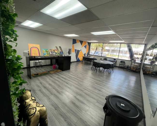 The image shows an empty office space with tables, chairs, artwork displayed on a counter, and large windows allowing natural light in.