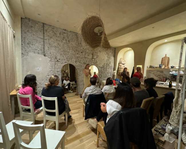 A group of people sits in a cozy, softly lit room with exposed brick walls, attentively watching a speaker stand in front of them.