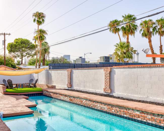 The image shows a swimming pool surrounded by palm trees and a wall, with lounge chairs on a grassy area adjacent to the pool.