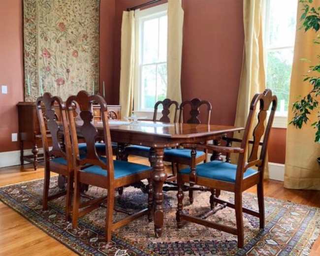 A wooden dining table with six chairs is arranged on a patterned rug in a room with brown walls and large windows.