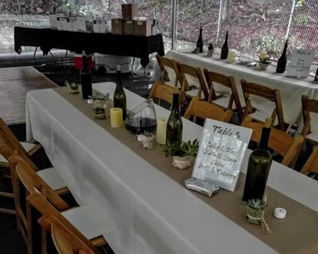 A long table is set for an event, featuring bottles, candles, and a sign indicating "Table 5," in a partially enclosed space.