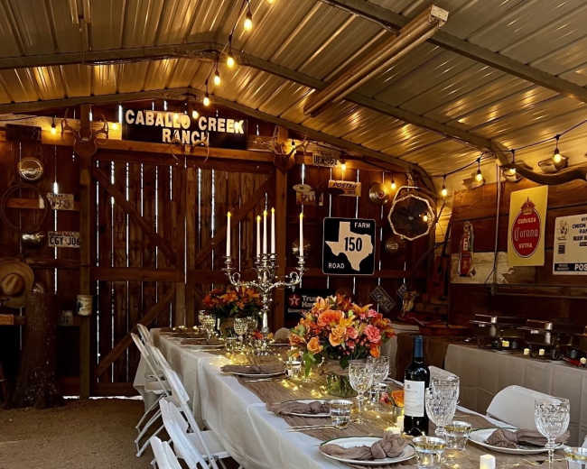 A long dining table is set with floral centerpieces and elegant tableware inside a rustic barn, adorned with string lights and vintage signs.