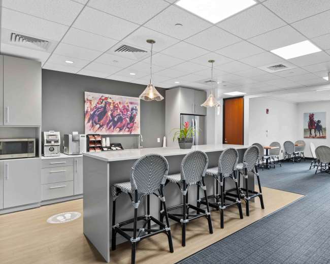 The image shows a modern office kitchenette with a bar counter, seating, and a dining area featuring artwork on the walls.