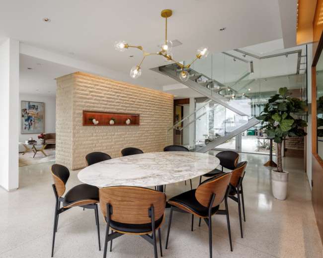 A modern dining area features a round marble table surrounded by black and wooden chairs, with a staircase and a textured stone wall in the background.