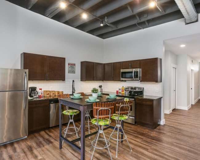 A modern kitchen features dark wooden cabinets, stainless steel appliances, and a central dining table with four stools.