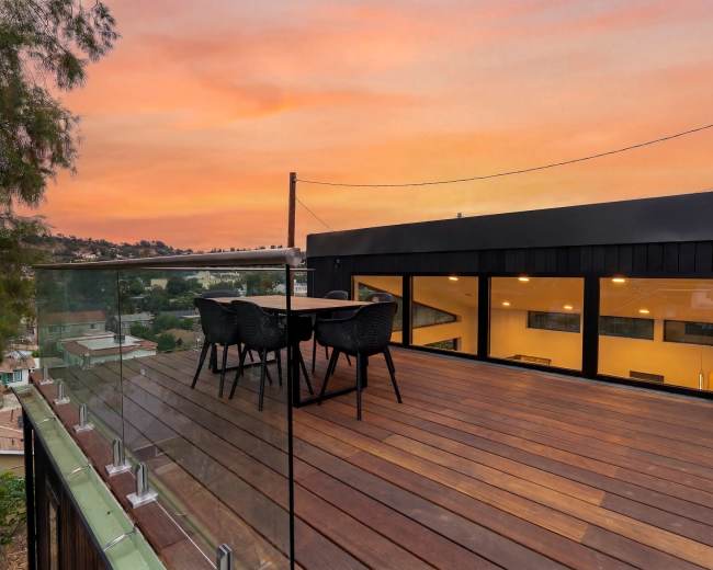 A rooftop deck with a wooden floor features a table and chairs, overlooking a scenic area at sunset.