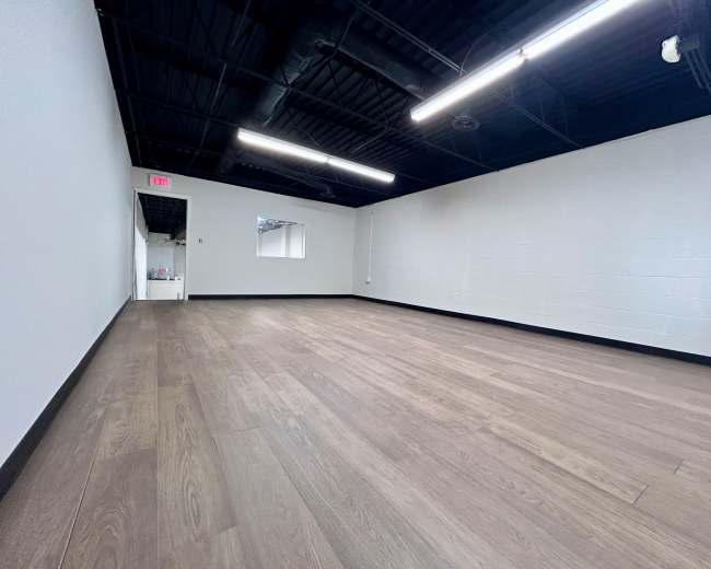 The image shows a spacious, empty room with wooden flooring, white walls, and a door leading to another area.