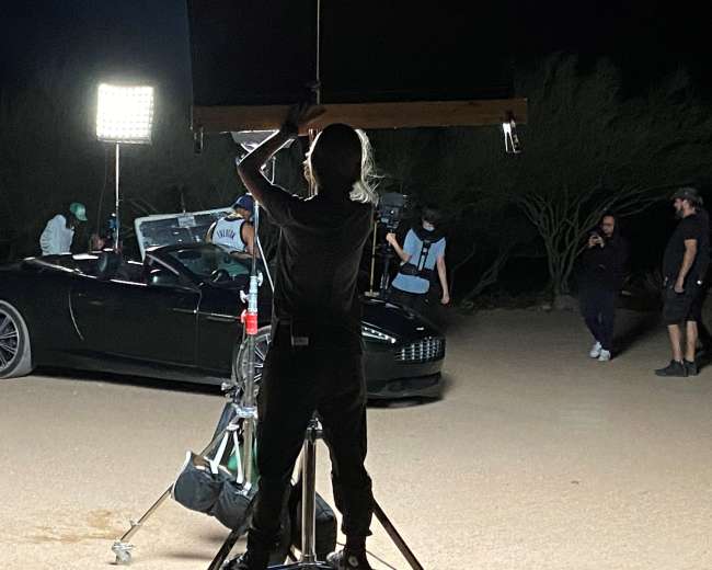 A crew member adjusts lighting equipment near a convertible car in a desert setting at night.