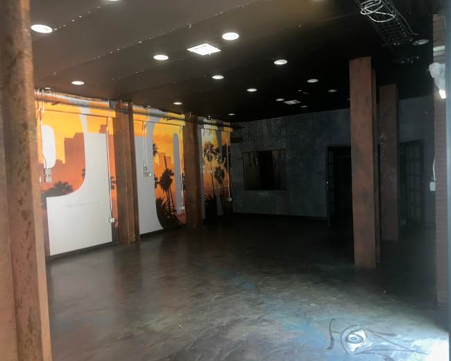 The image shows an industrial-style indoor space with dark flooring and exposed pillars, featuring a vibrant sunset mural on one wall.