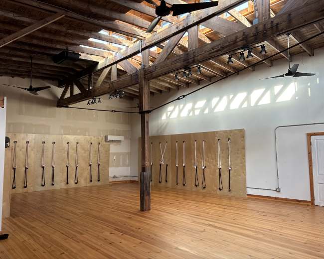The image shows a spacious room with wooden floors and high ceilings, featuring wooden beams and multiple fans, along with a wall displaying suspended straps for fitness activities.