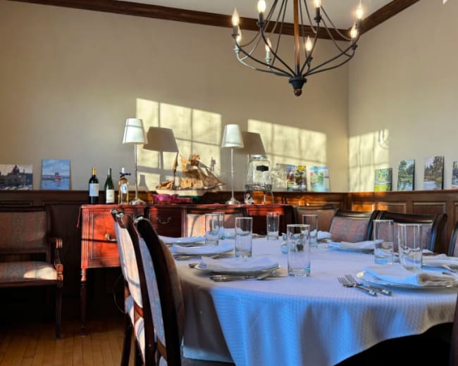A dining room with a large round table set for a meal, surrounded by wooden chairs, and decorated with various artworks, wine bottles, and a chandelier overhead.