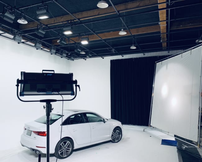 A white car is positioned in a well-lit studio with a black curtain in the background and lighting equipment visible in the foreground.