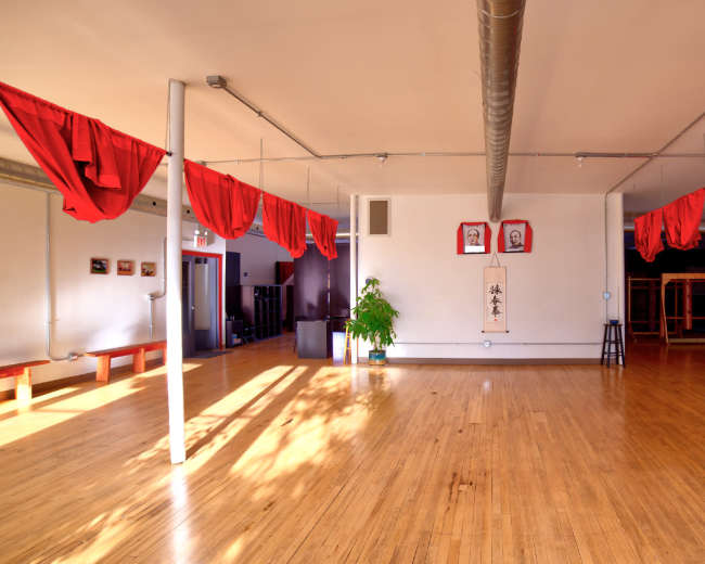 The image shows a spacious room with wooden floors, adorned with red fabric draped from the ceiling and featuring minimal decorations including framed art and a plant.