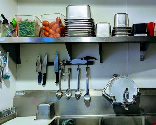 A kitchen workspace with metal shelving holding containers of vegetables, plates, and utensils, along with hanging knives and various tools near a stainless steel sink.