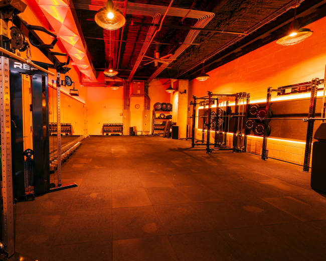 The image shows a well-lit gym with exercise equipment, including a cable machine and weight racks, set against a backdrop of orange lighting.