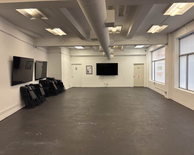 The image shows a spacious, empty room with several wall-mounted screens and a stack of black folding chairs in one corner.