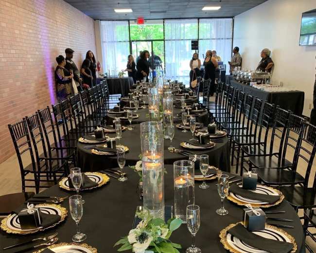 A long, elegantly set dining table with black tablecloths and gold accents is arranged for an event, featuring glasses, plates, and decorative centerpieces, while guests mingle in the background.