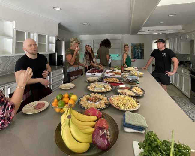 A group of people gathers around a kitchen island filled with various dishes and fruits.