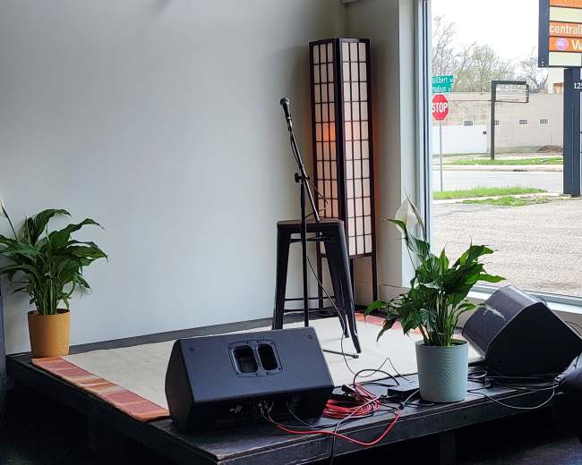 A small stage with a microphone and amplifier, flanked by potted plants, set in a room featuring large windows.