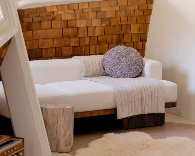 The image shows a cozy living space with a white couch, a round purple pillow, a wooden side table, and a soft rug on the floor.